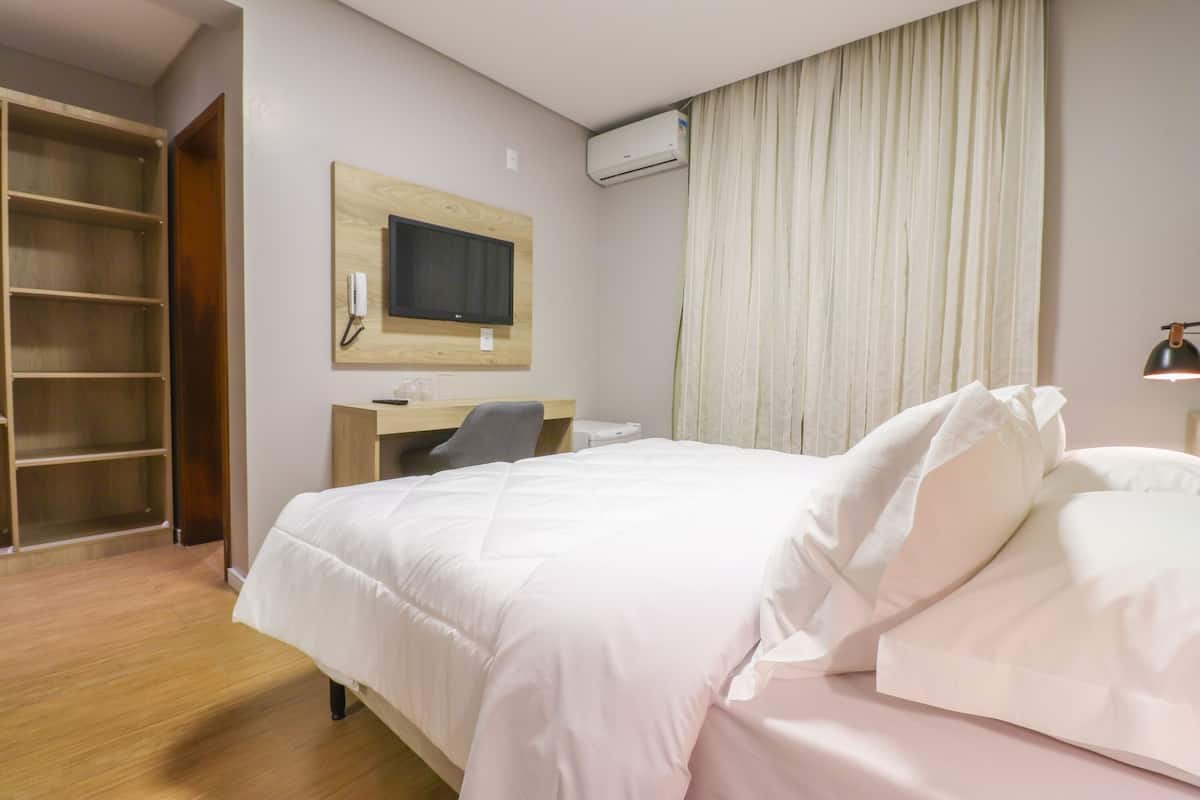 A hotel room with a bed, a desk, a chair, a wardrobe, and an air conditioning unit.