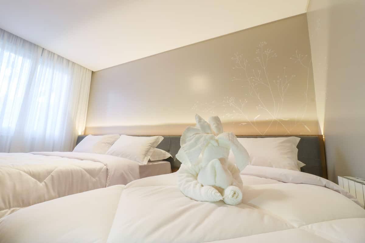 A hotel room with two beds, each with white bedding and pillows. There is a decorative towel arrangement on the bed in the foreground.