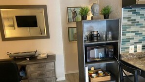 A compact kitchen with a microwave, a small refrigerator, and a coffee machine. There are two shelves with decorative items and a small table with a chair. - Treehouse Apartment Rentals (Pompano Beach)