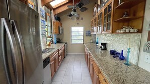 A kitchen with wooden cabinets, a stainless steel refrigerator, a granite countertop, and a window with blinds. - Treehouse Apartment Rentals (Pompano Beach)