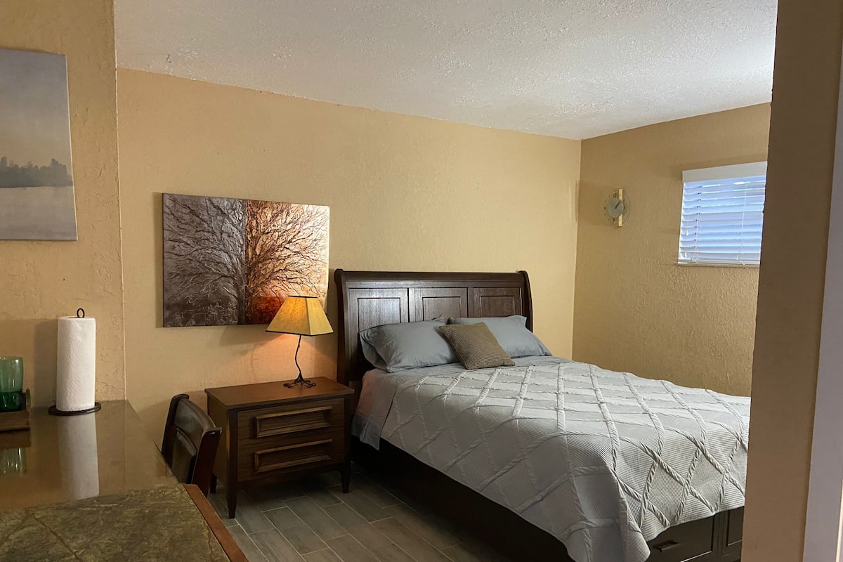 A bedroom with a bed, a nightstand, a lamp, a wall clock, and a window with blinds.