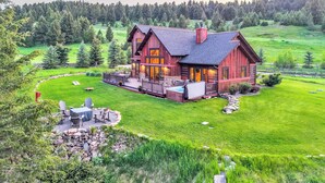Eine große, rustikale Holzhütte mit einer geräumigen Terrasse und einem Whirlpool, umgeben von üppiger grüner Landschaft und dichtem Wald.