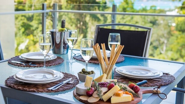 Mesa para dos personas con platos, vasos, una botella y una tabla de quesos con diversas variedades de quesos y frutas.