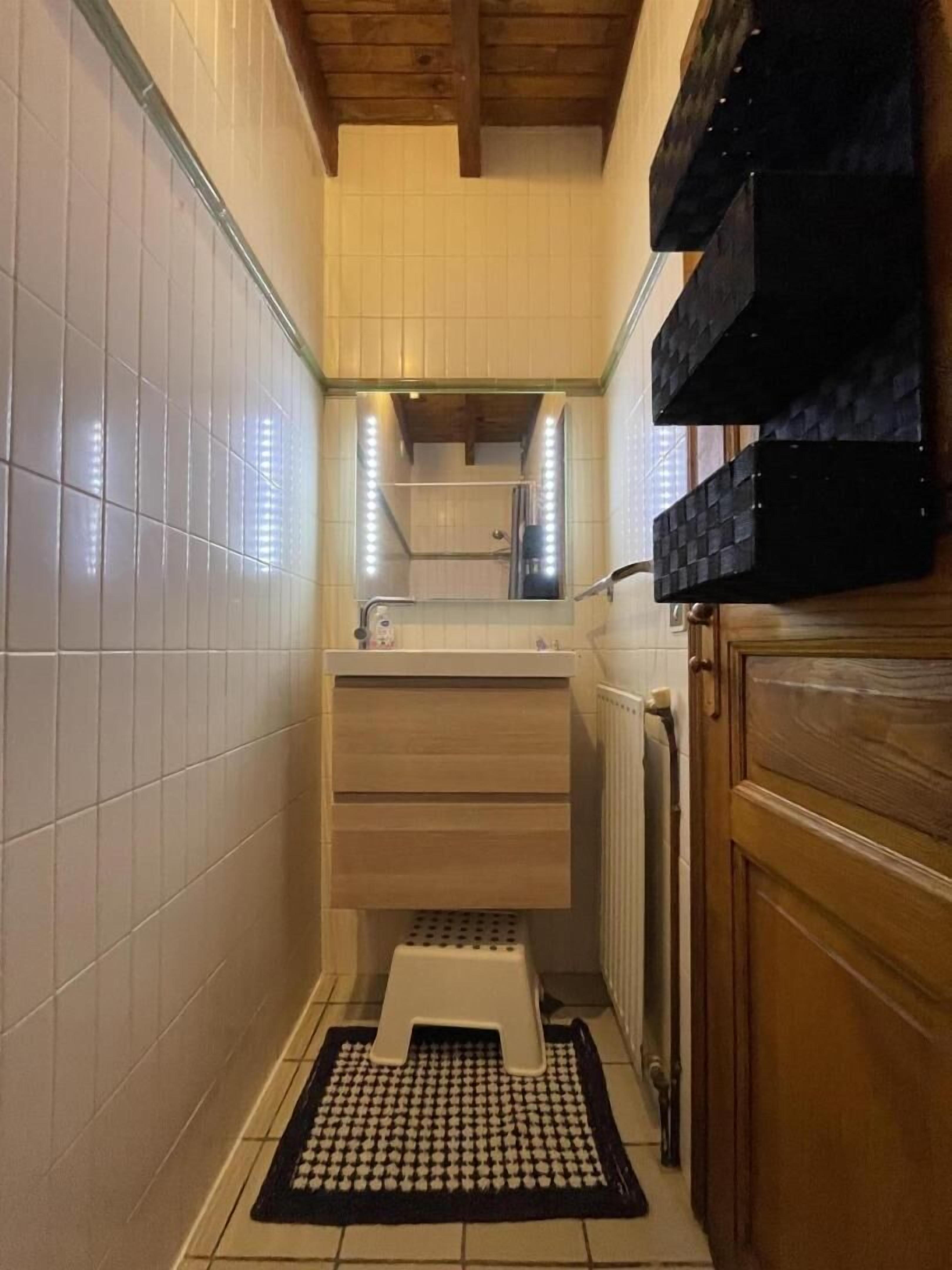 Een smalle badkamer met witte betegelde wanden, een houten plafond, een wastafel met spiegel en een zwarte mat op de vloer.