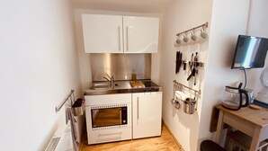 Een kleine keuken met witte kastjes, een inbouwoven, een kookplaat en een magnetron. Er hangen keukengerei aan de muur en een koffiezetapparaat staat op het aanrecht.