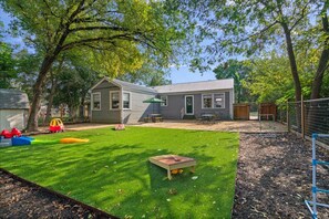 A backyard with a small house, a green lawn, a wooden play table, and a fenced play area.