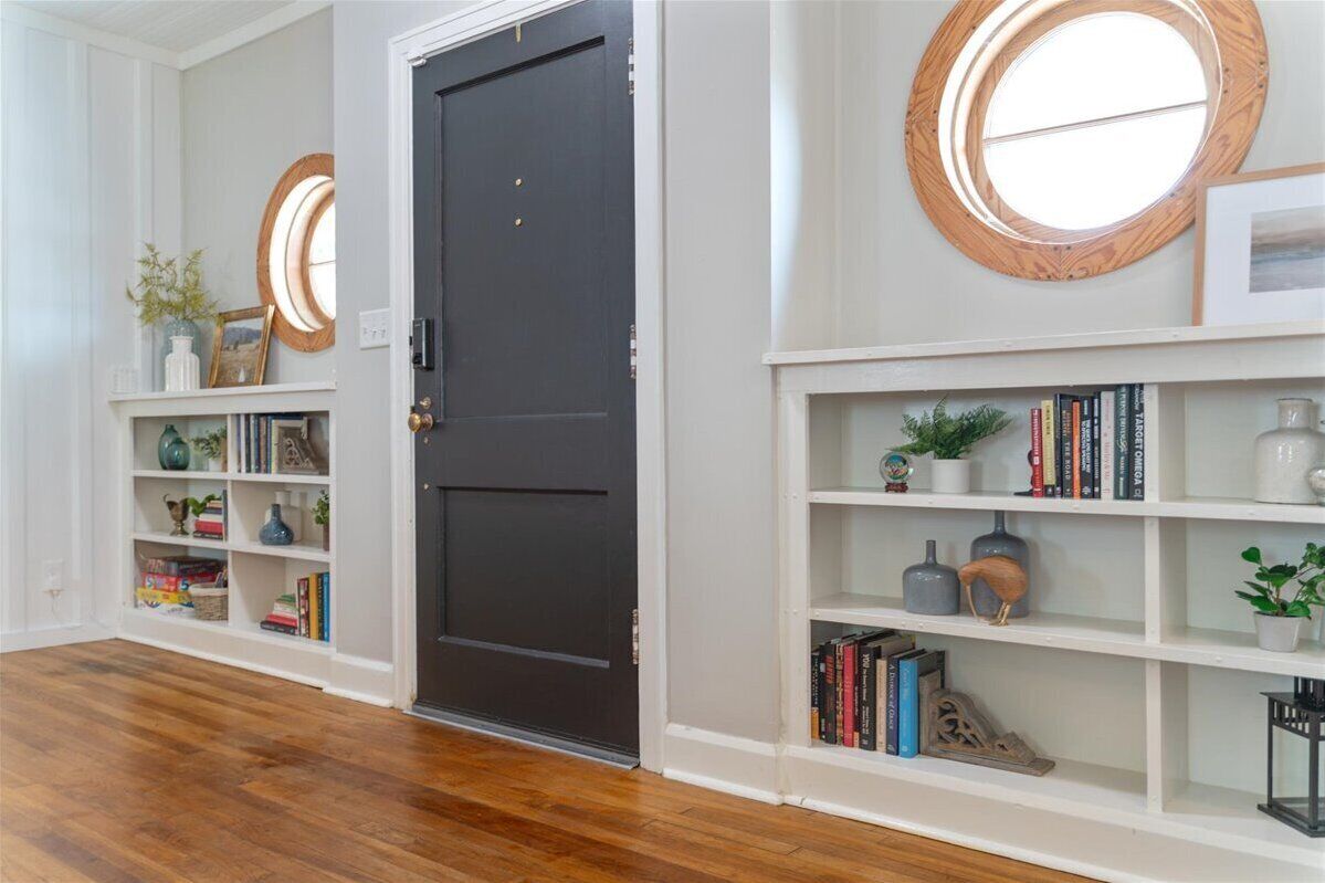 Un couloir avec une porte noire, des étagères blanches ornées de livres et d’objets décoratifs, et un miroir rond accroché au mur.