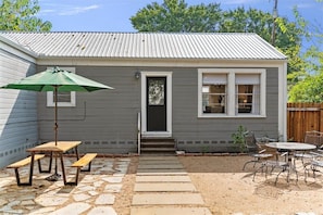 A small house with a gray exterior, a front door, a window, a patio with a table and chairs, and a green umbrella.