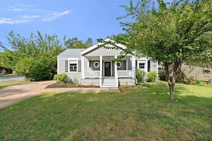 A single-story house with a front porch, a small front yard, and a tree to the right.