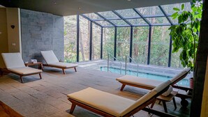 Un espace piscine intérieur moderne, doté de trois chaises longues, d’un mur en pierre et de grandes fenêtres offrant une vue sur la verdure environnante.