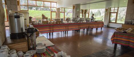 A spacious dining area with a long table set for a meal, surrounded by large windows offering a view of a garden.