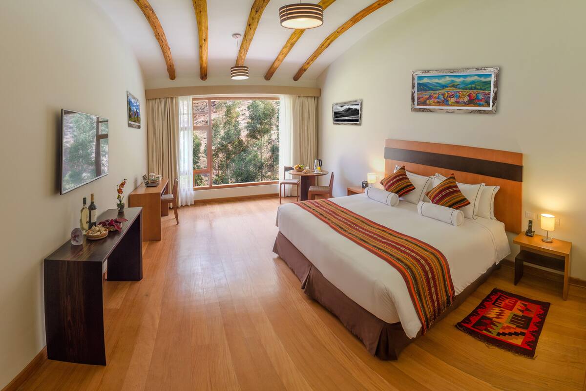 Une chambre spacieuse avec un grand lit, un bureau et une chaise. Il y a une fenêtre avec des rideaux, un tableau au mur et un plafond en bois.