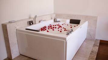 A large, rectangular bathtub filled with water and rose petals, with a handheld showerhead and a small control panel on the side.