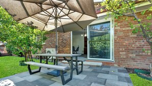 Une terrasse extérieure avec une table de pique-nique et un parasol, un mur en briques et une porte vitrée donnant sur une pièce.