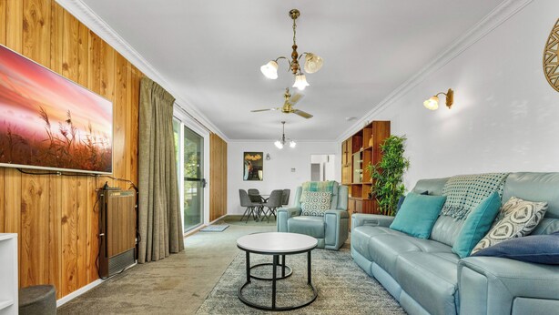 Un salon avec un canapé bleu, une table basse ronde et un ventilateur de plafond. On y trouve un mur en bois orné d’un tableau, une bibliothèque et une plante.