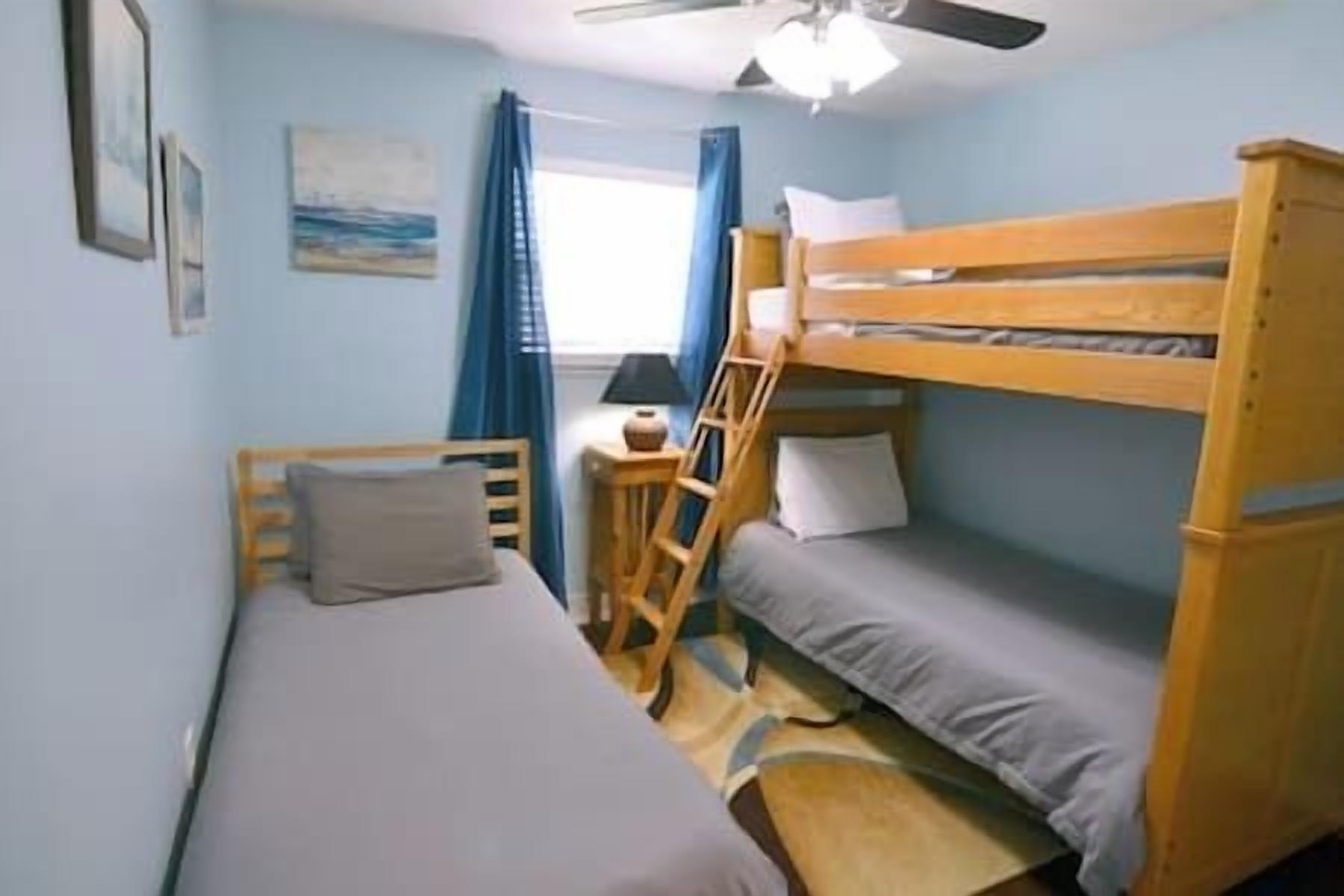 Une chambre avec un lit superposé, un lit simple, un bureau avec une chaise, une fenêtre avec des rideaux et un ventilateur de plafond.