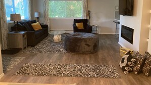 A living room with a sofa, ottoman, and coffee table, a TV mounted on the wall, and a window with curtains. - Downtown at your Doorstep (Anchorage)