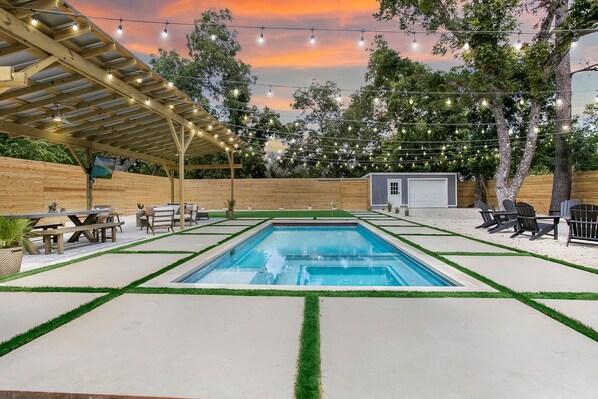Un jardin avec une piscine rectangulaire, entourée d’une terrasse en bois et d’un espace patio couvert.