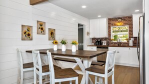 Una cocina con armarios blancos, una mesa de comedor de madera y sillas, con un cuadro de girasoles en la pared.