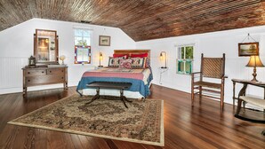 A bedroom with a sloped ceiling, wooden floor, a bed with a patterned bedspread, a bench, a dresser, a chair, and a table with a lamp. - 3/2 Historic Home | Walk to Main | Full Kitchen | WiFi | Hot Tub | Children over 10 welcome! (Fredericksburg)