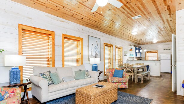 Un salón acogedor con sofá, reposapiés y sillas coloridas, techo de madera y una zona de comedor con mesa y sillas.