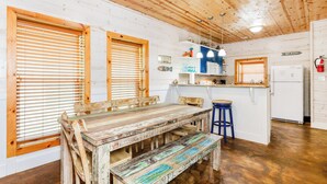 Interior rústico de una cabaña con una mesa de madera, bancos, una zona de cocina y un letrero que indica la dirección a la playa.