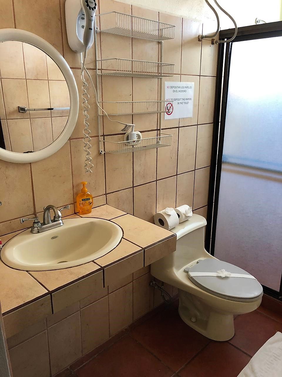 Ein Badezimmer mit Waschbecken, Toilette und Spiegel. An der Wand hängt ein Schild und auf der Ablage steht eine Flasche Handseife.