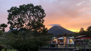 A parking lot with a building in the background, a tree in the foreground, and a mountain in the distance. - Apartment 10: Condos Vista al Volcan - Second Floor (La Fortuna)