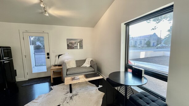 A modern living room with a grey sofa, a small round table, a white rug, and a large window offering a view of the street. - The No. 3: modern, bright, renovated, dwtn condo w/ outdoor lounge space (Marfa)