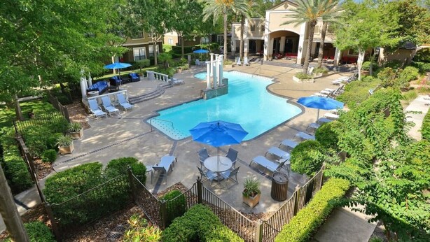 A pool area with lounge chairs and umbrellas, surrounded by greenery and buildings. - Large 1 Bedroom Apartment on Westheimer Road (Houston)