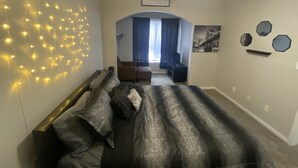 A bedroom with a bed, a ceiling fan, string lights, a window with curtains, and a cityscape painting. - Large 1 Bedroom Apartment on Westheimer Road (Houston)