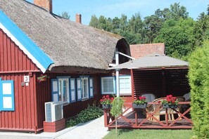 A rustic wooden building with a thatched roof, a covered outdoor seating area with wicker furniture, and a small garden with potted plants. - Amber Retreat Nida with Free Parking & 2 Bedrooms  (Neringa)
