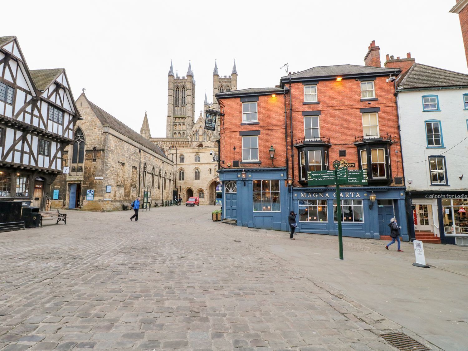 A cobblestone square with historic buildings, including a pub named 'Magna Carta' and a church with a tall spire in the background.