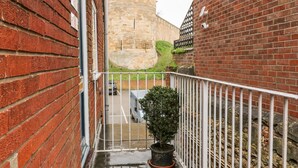 A narrow alleyway with a brick wall on the left and a metal railing on the right. A potted plant is in the foreground, and a stone tower is visible in the distance. - Grayz Studio (Lincoln)