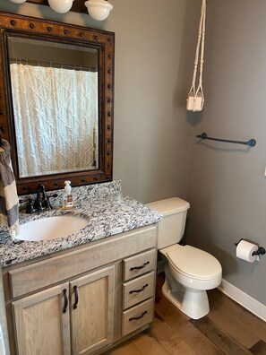 A bathroom with a granite countertop sink, a wooden vanity with drawers, a white toilet, and a mirror with a decorative frame. - McCrary Clan Lodge (Counce)