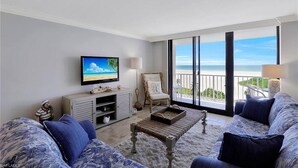 Una sala de estar con televisor, un sillón de mimbre, un sofá y un balcón con vista al océano.
