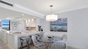 Cocina moderna con muebles blancos, una isla central y un comedor con sillas de mimbre y una mesa de vidrio.