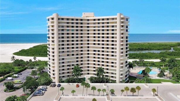 Un hotel de varios pisos con fachada curva, situado cerca de una playa con piscina y palmeras.