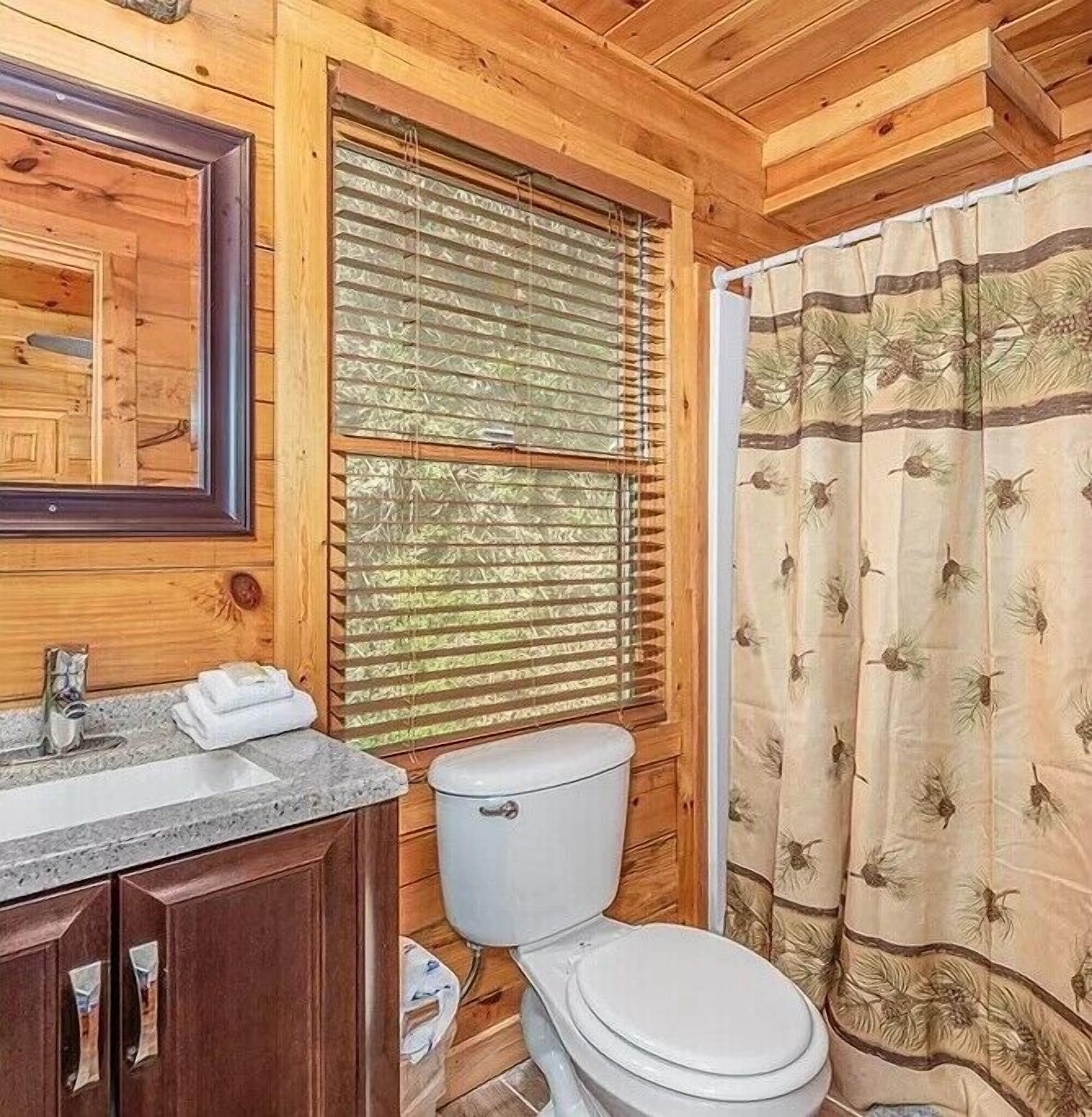 A wooden cabin bathroom with a sink, mirror, toilet, and window with blinds.