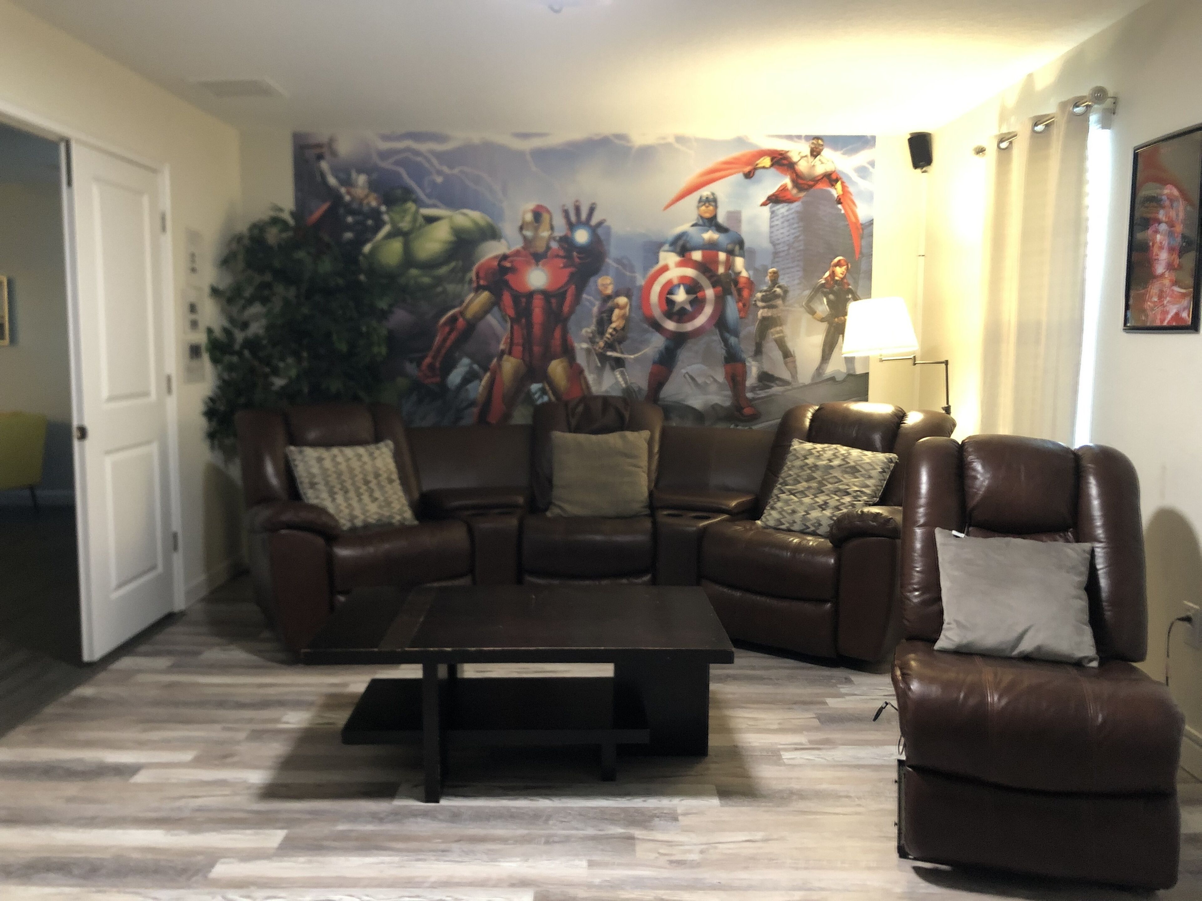 Een woonkamer met een bruine leren zithoek, een salontafel en een grote muurschildering met superhelden.