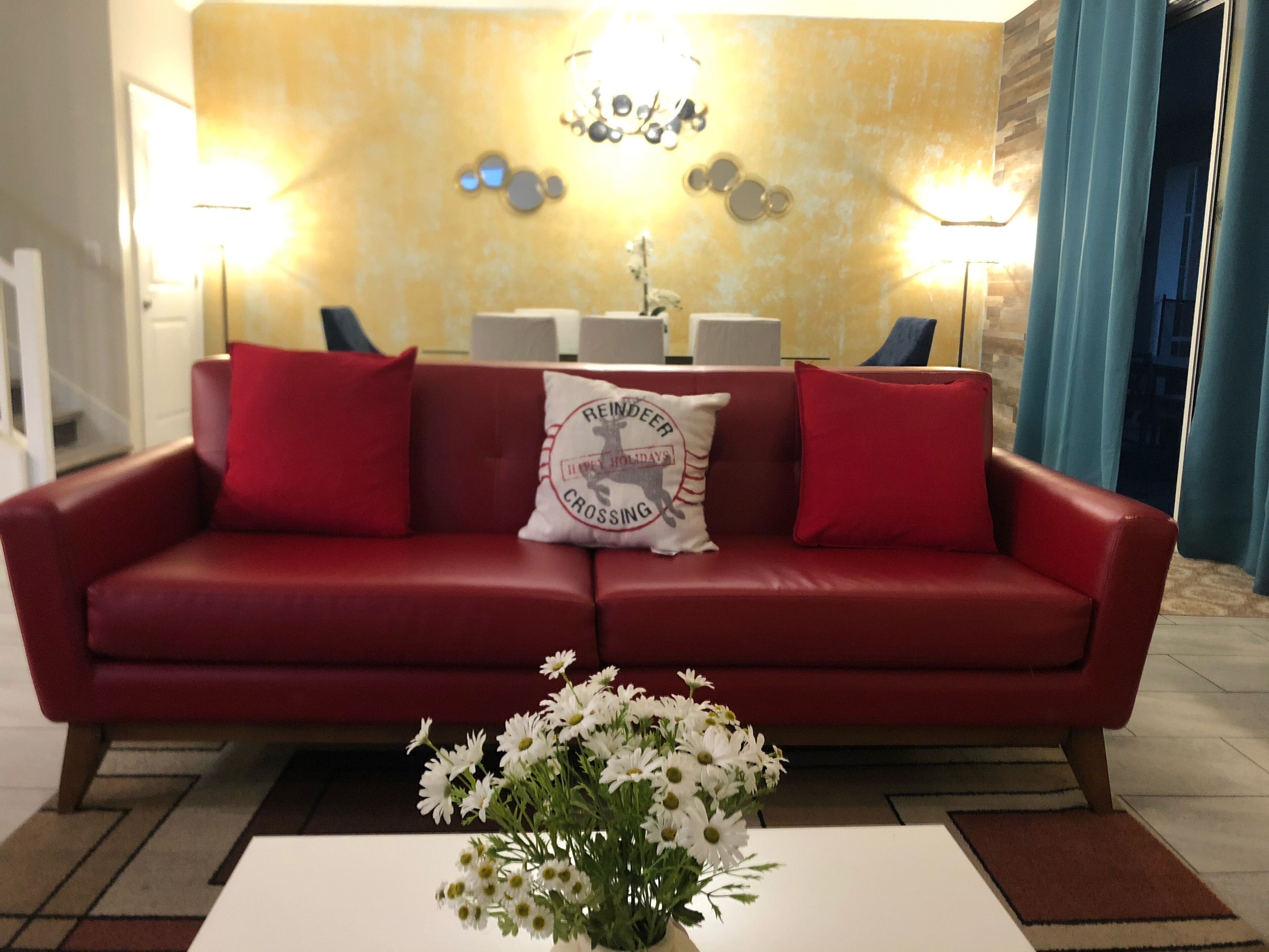 A red leather sofa with two red cushions and a white pillow with a logo. In front of the sofa is a white table with a vase of white flowers.