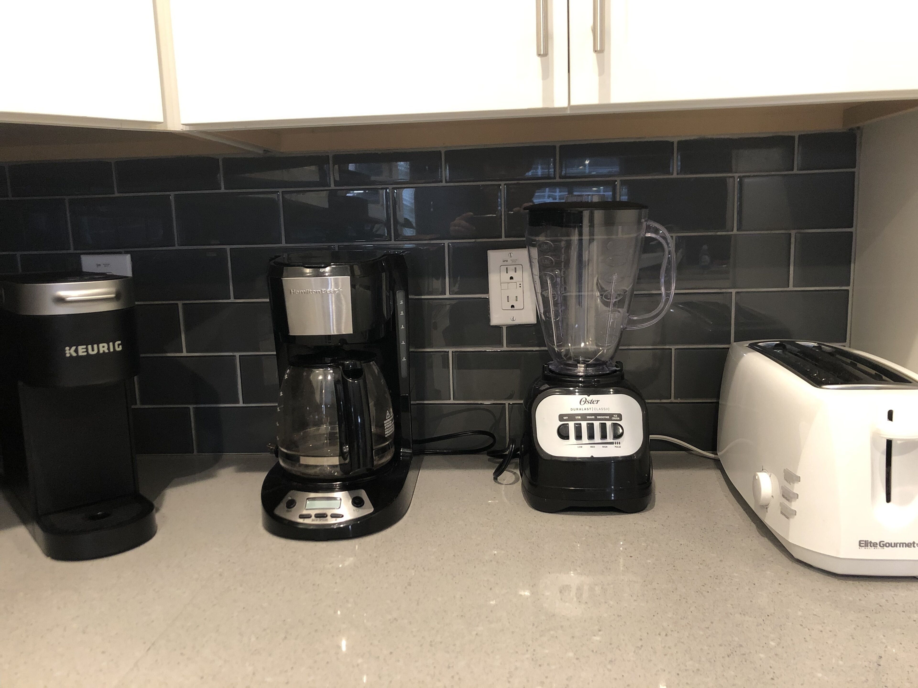 Uma bancada de cozinha com uma cafeteira Keurig, um liquidificador Hamilton Beach e uma torradeira Oster.