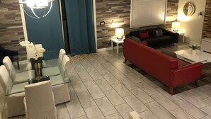 Ett modernt vardagsrum med ett soffbord i glas, en röd soffa, en svart soffa och en vit fåtölj. Det finns en blå gardin, en vägglampa och en takfläkt.