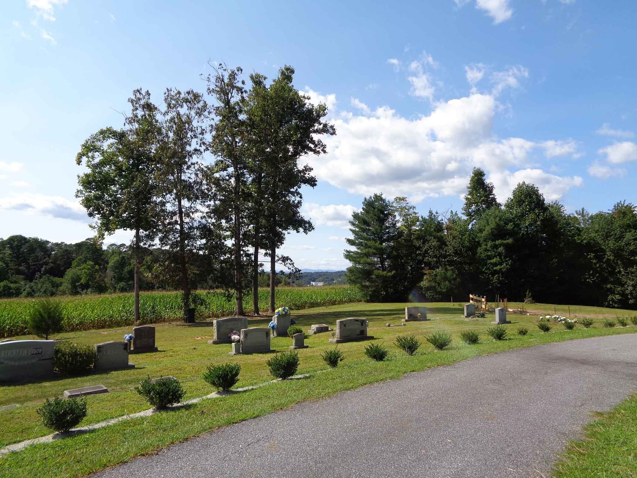 Un cimetière avec de nombreuses pierres tombales, un chemin pavé et une zone herbeuse parsemée d’arbres.
