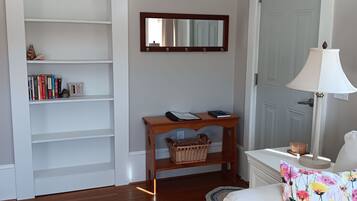 A living room with a wooden floor, a white sofa, a small table with a lamp, a bookshelf, and a door.