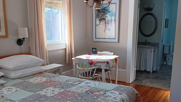 Ein Schlafzimmer mit einem großen Bett, einem Nachttisch, einem Sessel, einem Tisch mit floralem Tischtuch, einer Deckenleuchte, einem Fenster mit Vorhängen, einem Gemälde und Blick ins Badezimmer.