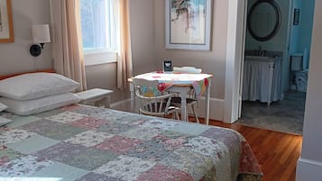 A bedroom with a large bed, a bedside table, a chair, a table with a floral tablecloth, a chandelier, a window with curtains, a painting, and a view of the bathroom.