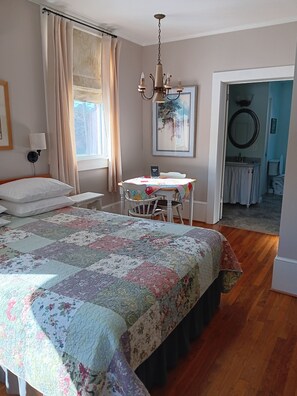 Een slaapkamer met een groot bed, een nachtkastje, een stoel, een tafel met bloemendessin, een kroonluchter, een raam met gordijnen, een schilderij en uitzicht op de badkamer.