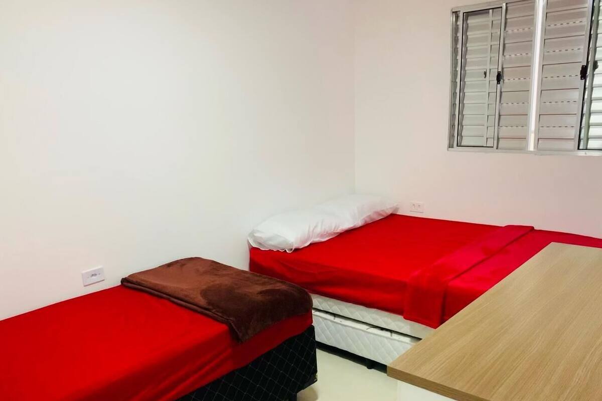 Una habitación pequeña y limpia con dos camas individuales, una mesa de madera y una ventana con persianas.
