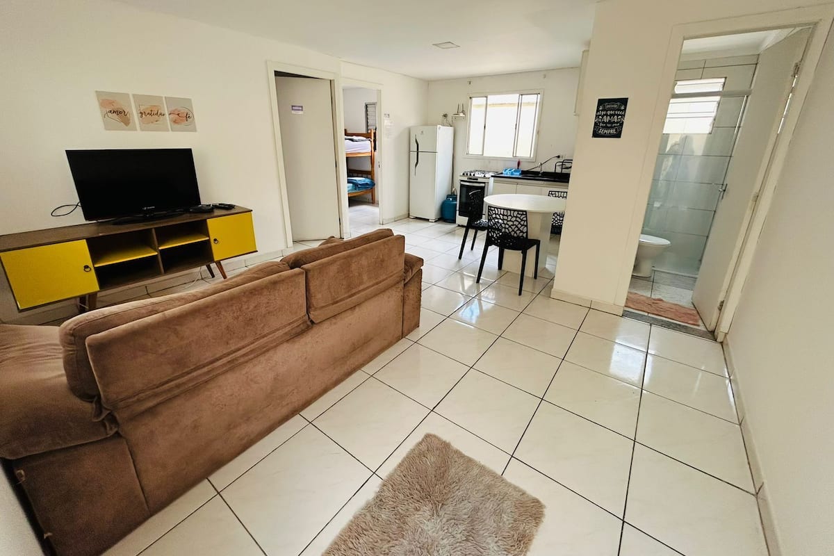 Sala de estar con sofá, televisor sobre un mueble amarillo, área de cocina con mesa y sillas, y un baño visible a través de un umbral abierto.